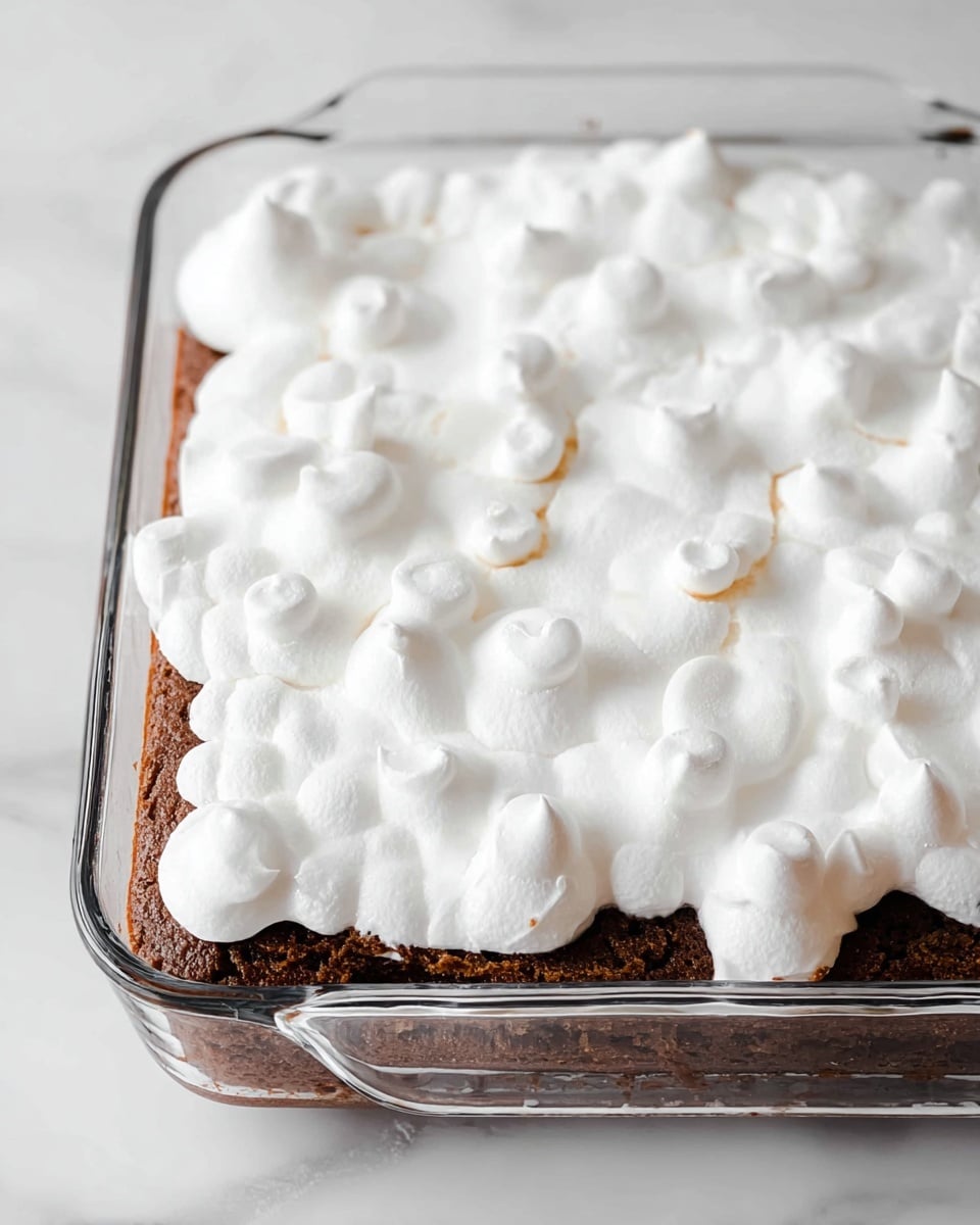 The image shows a clear glass baking dish containing a dessert with two visible layers. The bottom layer is a dark brown cake with a soft, slightly crumbly texture. On top is a thick, fluffy white layer of marshmallow or meringue spread unevenly, with small rounded peaks and some thin spots revealing the cake beneath. The dish is placed on a white marbled surface. photo taken with an iphone --ar 4:5 --v 7