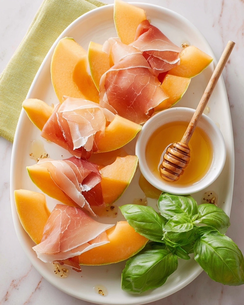 A white oval plate holds eight slices of bright orange cantaloupe, each wrapped loosely in thin, light pink slices of prosciutto with white fat streaks. Near the top right of the plate is a small white bowl filled with golden honey, with a wooden honey dipper resting inside it. To the right side of the plate, fresh bright green basil leaves add a pop of color. The plate is set on a white marbled surface that contrasts softly with the colors of the food. Photo taken with an iphone --ar 4:5 --v 7