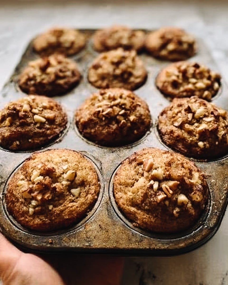 The image shows a close-up of a muffin tray filled with freshly baked muffins. Each muffin has a golden brown top with a rough texture, covered with small pieces of nuts which add a crunchy look. The muffins are slightly risen with a soft dome shape and evenly spaced in the metal muffin tray. The tray sits on a white marbled surface, and a woman’s hand is holding the edge of the tray from the right side of the frame. photo taken with an iphone --ar 4:5 --v 7