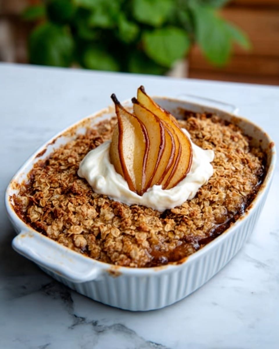 The image shows a white baking dish filled with a baked fruit crisp with a crumbly, golden-brown oat topping covering the whole surface. In the center, there is a dollop of white cream topped with several thin slices of caramelized brown pear arranged in a small stack. The dish is placed on a white marbled surface with a blurred green plant in the background. photo taken with an iphone --ar 4:5 --v 7