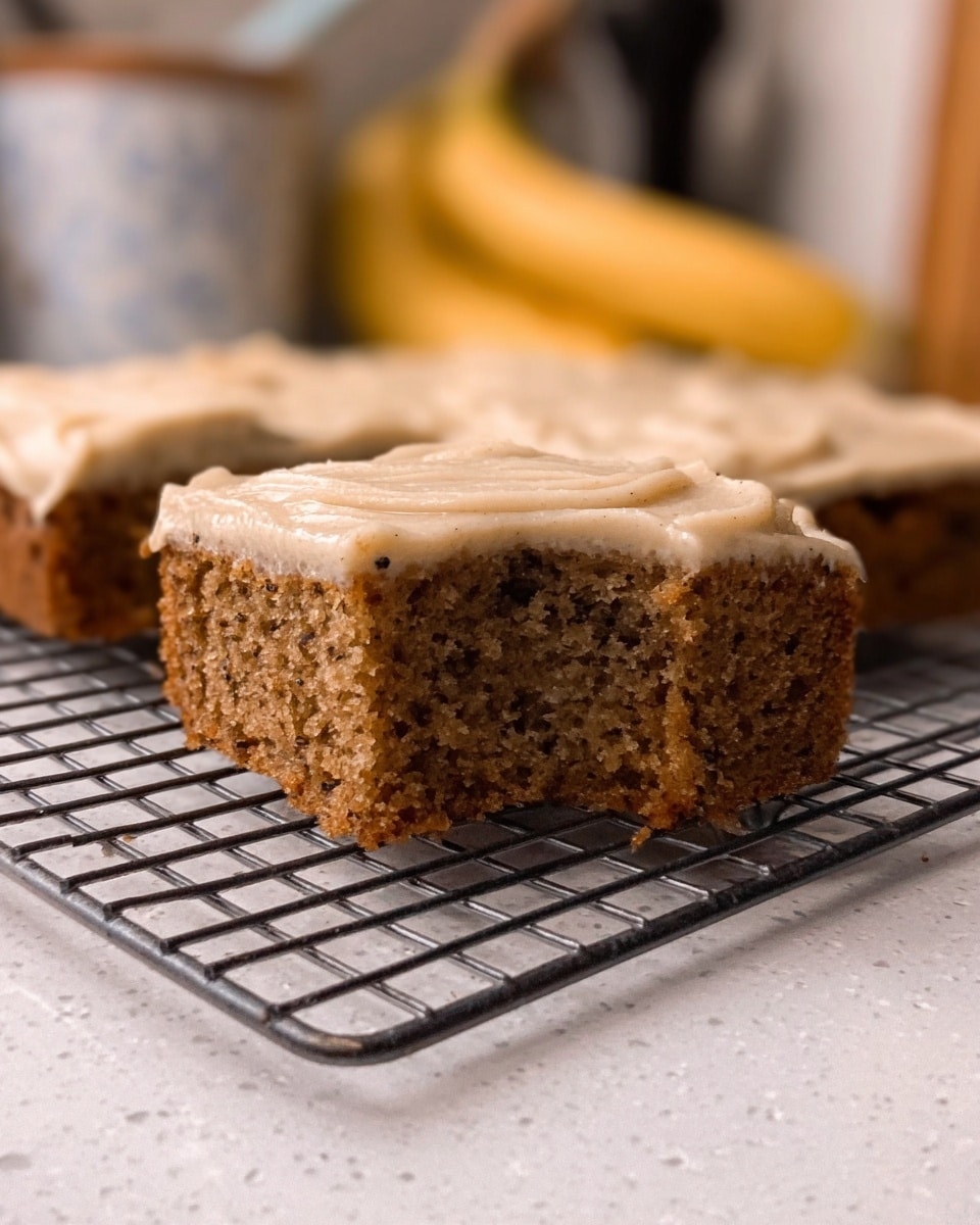 A close-up of a square brown cake with visible small bits inside, topped with a thick, light beige frosting spread evenly. One piece is lifted, showing the moist texture inside, while the rest of the cake remains on a metal cooling rack. The background features a white marbled surface, with some kitchen items and bananas blurred behind. Photo taken with an iphone --ar 4:5 --v 7