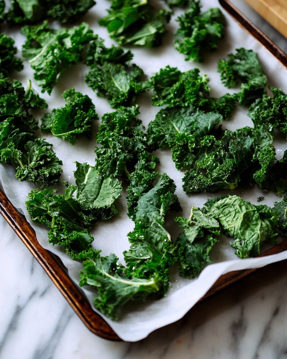 A baking tray with a layer of white parchment paper holds evenly spread pieces of fresh kale leaves, about three dozen, each dark green with a mix of smooth and crinkled textures, showing veins and curly edges. The tray is placed on a white marbled surface, and there is soft natural light illuminating the scene, emphasizing the fresh, vibrant green color and slight gloss on the leaves. In the background, the blurred edge of a box and some indistinct objects add depth to the image. Photo taken with an iphone --ar 4:5 --v 7