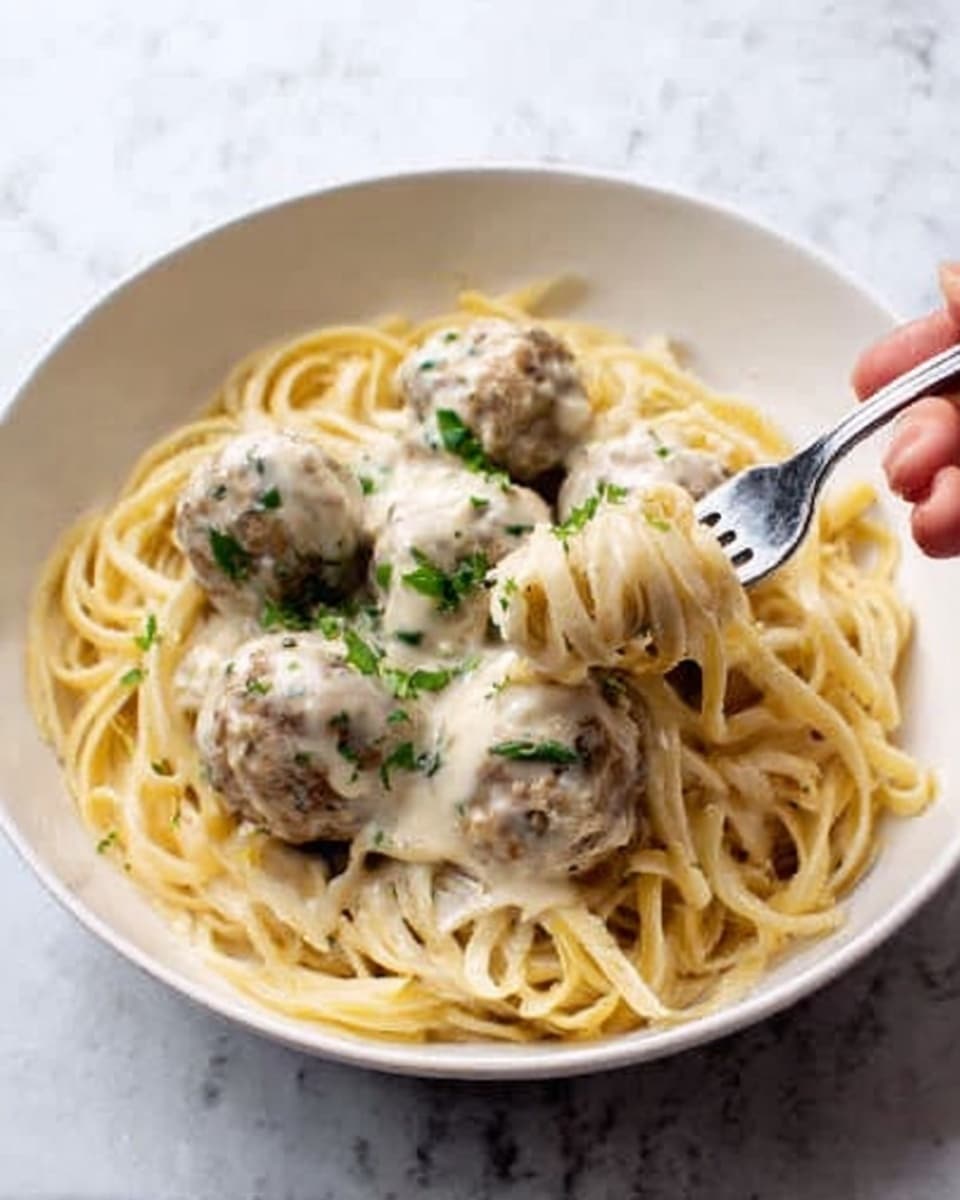 A white bowl filled with a bed of light yellow spaghetti noodles at the bottom, topped with several light brown meatballs covered in a thick, creamy white sauce mixed with small green herb pieces. A fork held by a woman's hand is lifting some noodles and meatballs from the bowl. The background is a white marbled texture. photo taken with an iphone --ar 4:5 --v 7