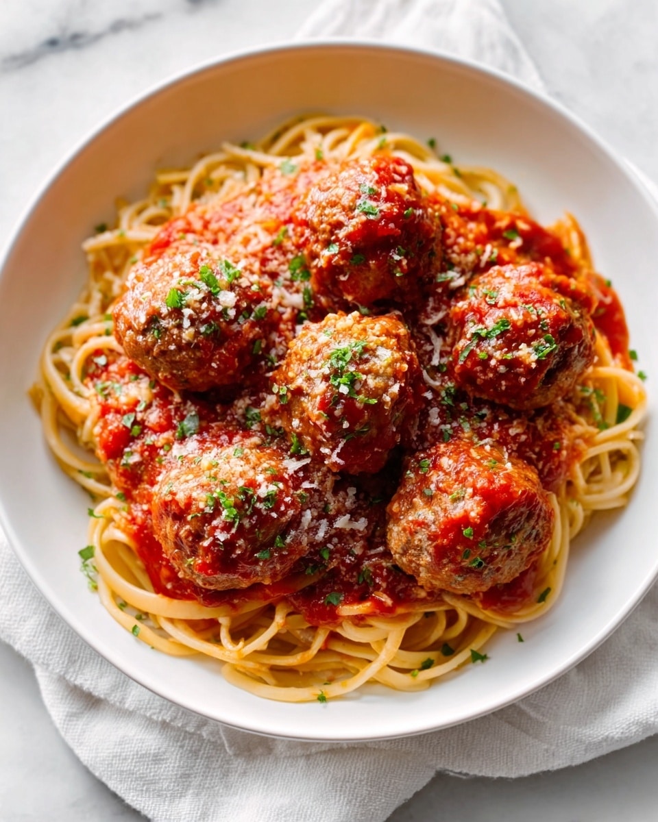 A white plate holds a nest of thin spaghetti noodles topped with five round meatballs covered in red tomato sauce. The sauce glistens and coats the pasta and meatballs evenly. Grated cheese and small green herb pieces are sprinkled on top of the meatballs and sauce. The plate is on a white marbled surface with a white cloth napkin slightly visible beneath. Photo taken with an iphone --ar 4:5 --v 7