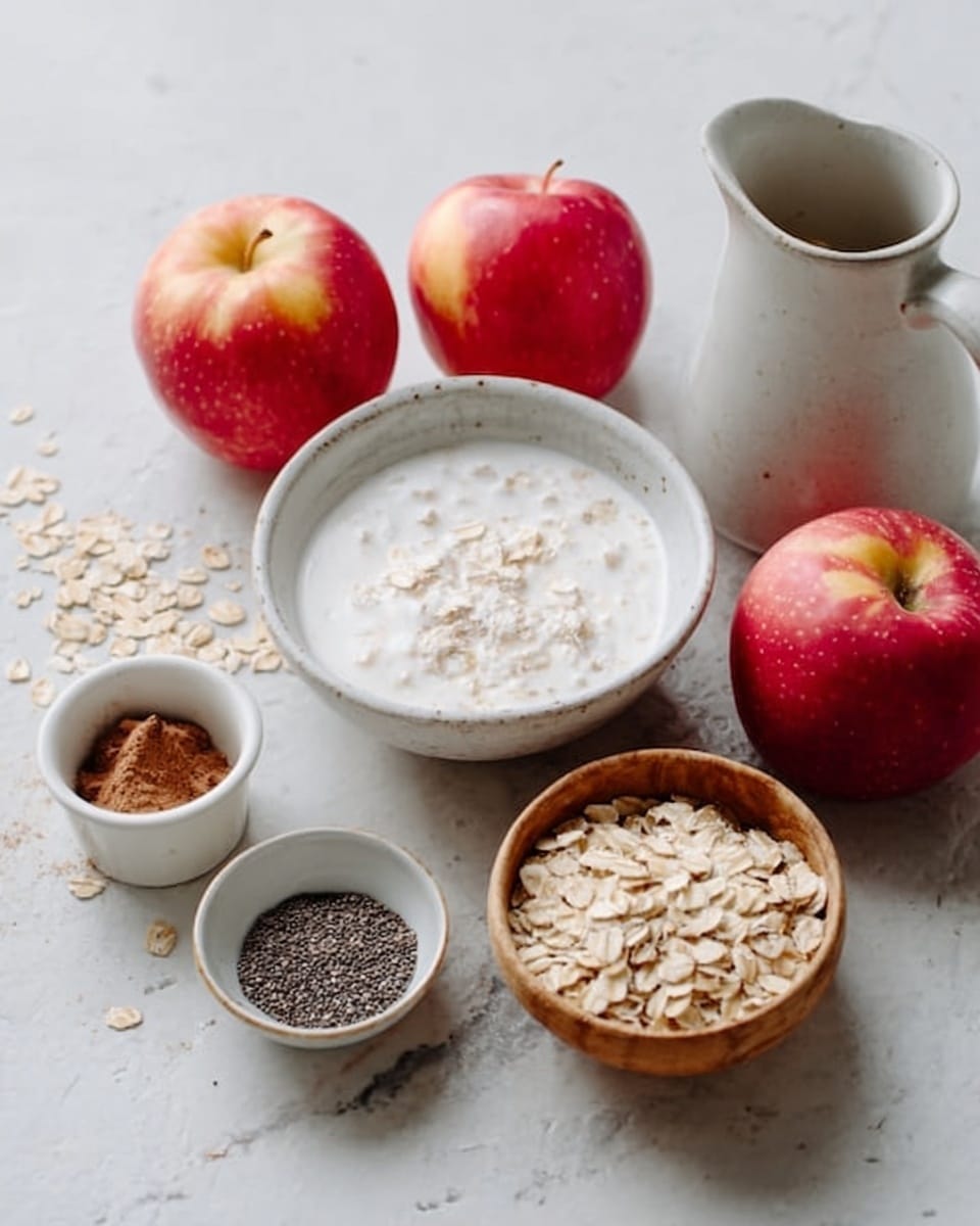 A white bowl filled with creamy white milk topped with white oat flakes sits on a white marbled surface. Nearby, a wooden bowl holds light brown rolled oats, with some oats scattered around it. To one side, there is a small white cup with a brown powder, and another small white cup filled with black chia seeds. Several small red apples with smooth skin are arranged around the bowls and cups. A white jug with a smooth surface sits in the background. Photo taken with an iphone --ar 4:5 --v 7