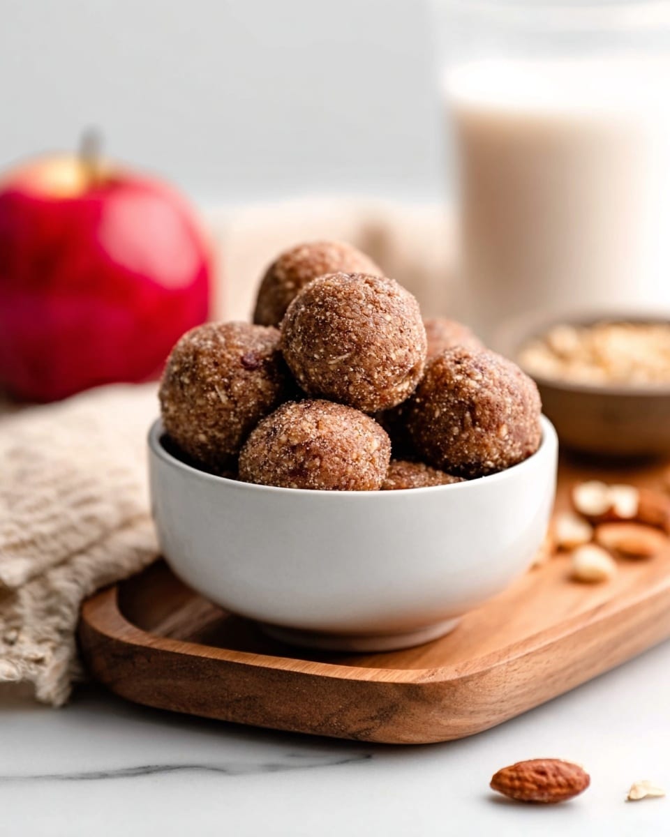 A white bowl filled with round, brown energy balls that have a slightly rough texture. The bowl is placed on a wooden board, which sits on a white marbled surface. In the background, there is a red apple on the left, a glass of milk on the right, and a small bowl of nuts nearby, all slightly blurred. The lighting is soft and natural, highlighting the texture of the energy balls. photo taken with an iphone --ar 4:5 --v 7