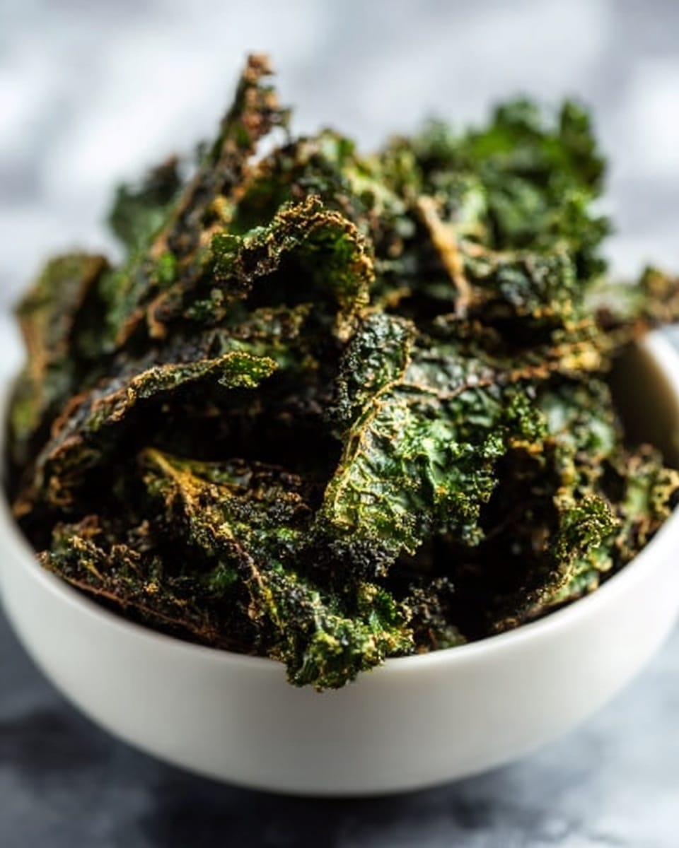 A round white bowl filled with several crispy kale chips showing deep green and slightly brown colors, with a rough and crunchy texture. The kale pieces are stacked loosely inside the bowl, filling it near the top. The background is a white marbled surface, softly blurred to keep focus on the kale chips. photo taken with an iphone --ar 4:5 --v 7