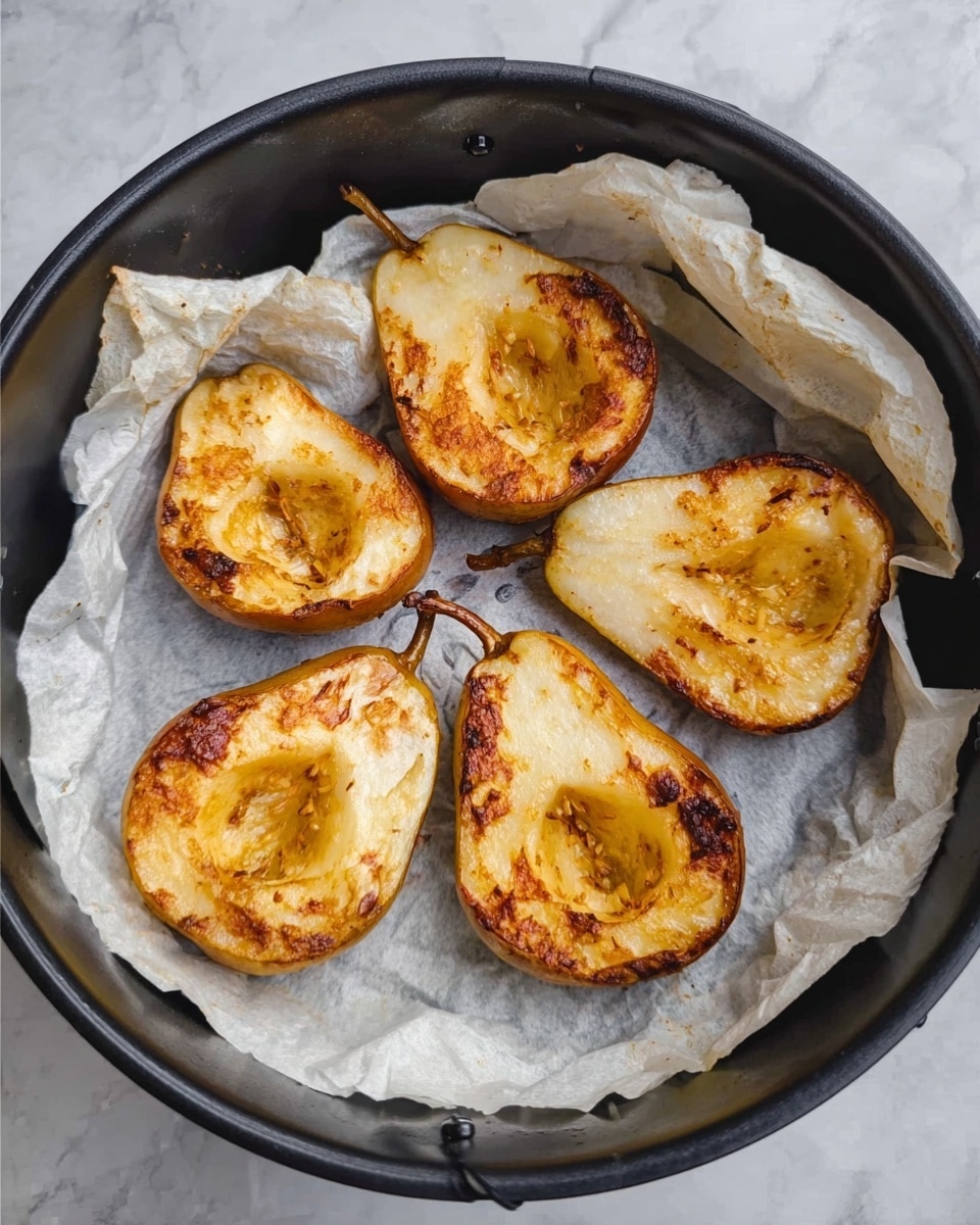The image shows four baked pear halves arranged in a circular form on white baking paper, inside a black round air fryer basket. The pears are light golden yellow with a caramelized brown center and edges, showing soft textures with slight wrinkling. The white marbled surface is visible around the basket. Photo taken with an iphone --ar 4:5 --v 7