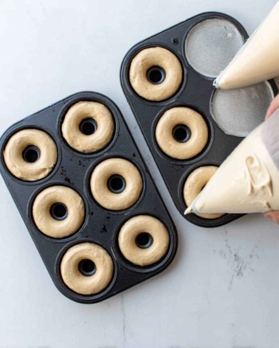The image shows two black donut pans, each holding five raw donut doughs. The doughs are light beige, smooth, and cylindrical with a large hole in the center, filling each mold evenly. To the right side, there is a large piping bag with cream-colored donut batter. The whole setup is placed on a white marbled surface, and a woman's hand can be seen holding the piping bag, ready to fill more molds. The scene is bright and clean, highlighting the smooth textures of the doughs and the neat arrangement. photo taken with an iphone --ar 4:5 --v 7