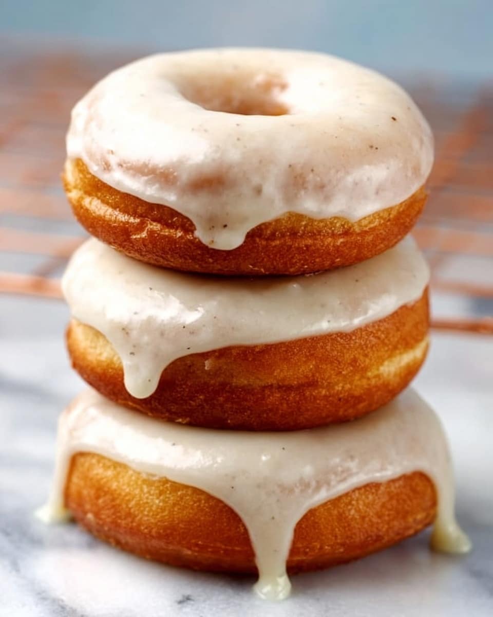 Three soft, golden brown donuts are stacked vertically on a white marbled surface, each topped with a thick layer of smooth white icing that drips slightly down the sides. The icing looks shiny and creamy with a few small specks visible on top, suggesting a subtle flavor detail. The bottom donut shows the icing flowing more freely over its side, while the middle and top donuts have a more even coat. The background is softly blurred to keep the focus on the donut stack. photo taken with an iphone --ar 4:5 --v 7
