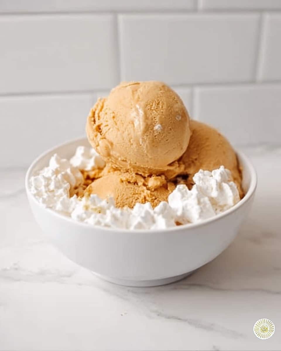 A white bowl sits on a white marbled surface with two large, round scoops of light brown ice cream in the center, surrounded by fluffy white whipped cream dollops evenly spaced around the edges of the scoops. The background shows a white tiled wall with a clean, bright atmosphere. The ice cream texture looks smooth yet slightly firm, and the whipped cream adds a soft, airy contrast. photo taken with an iphone --ar 4:5 --v 7