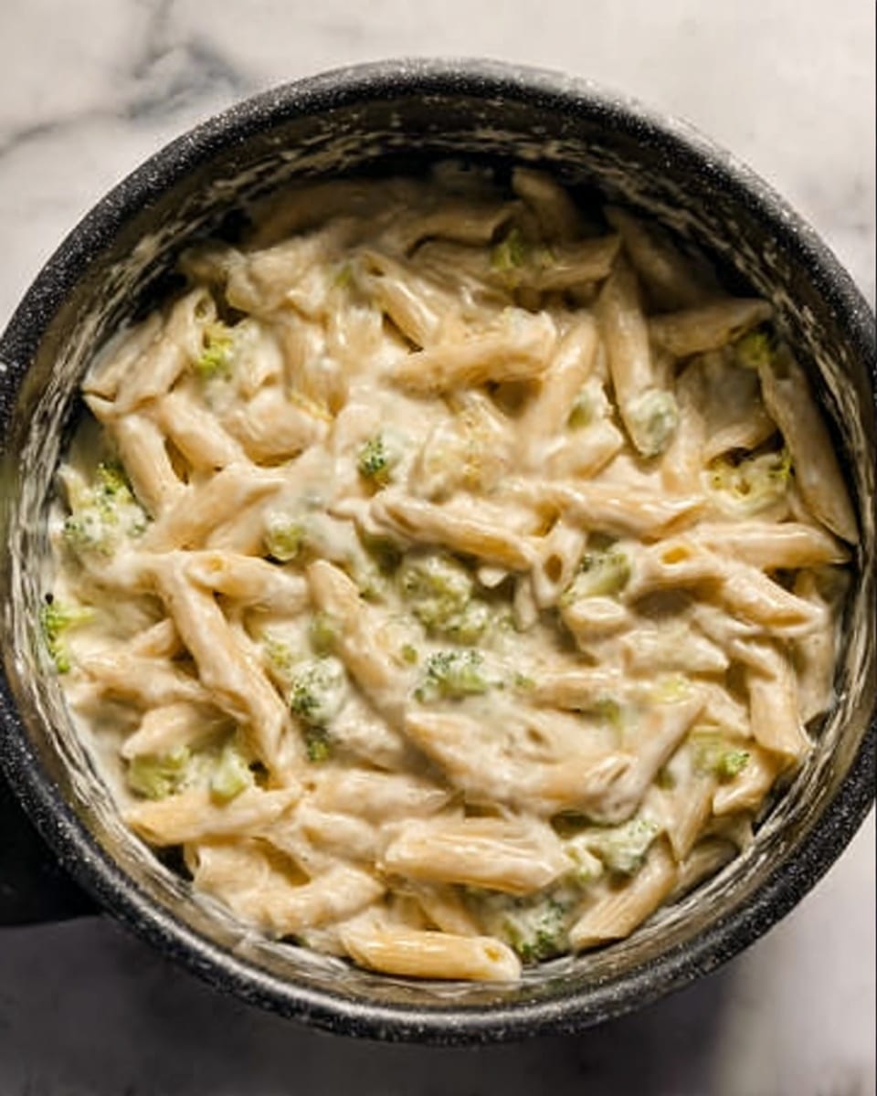 The image shows a black pot filled with creamy penne pasta mixed with small pieces of light green broccoli. The sauce is thick and white, covering the pasta evenly, giving a smooth texture. The pasta is soft and slightly shiny, while the broccoli adds a bit of rough texture and color contrast. The inside of the pot has some white sauce splashes. The background surface is white marble. Photo taken with an iphone --ar 4:5 --v 7