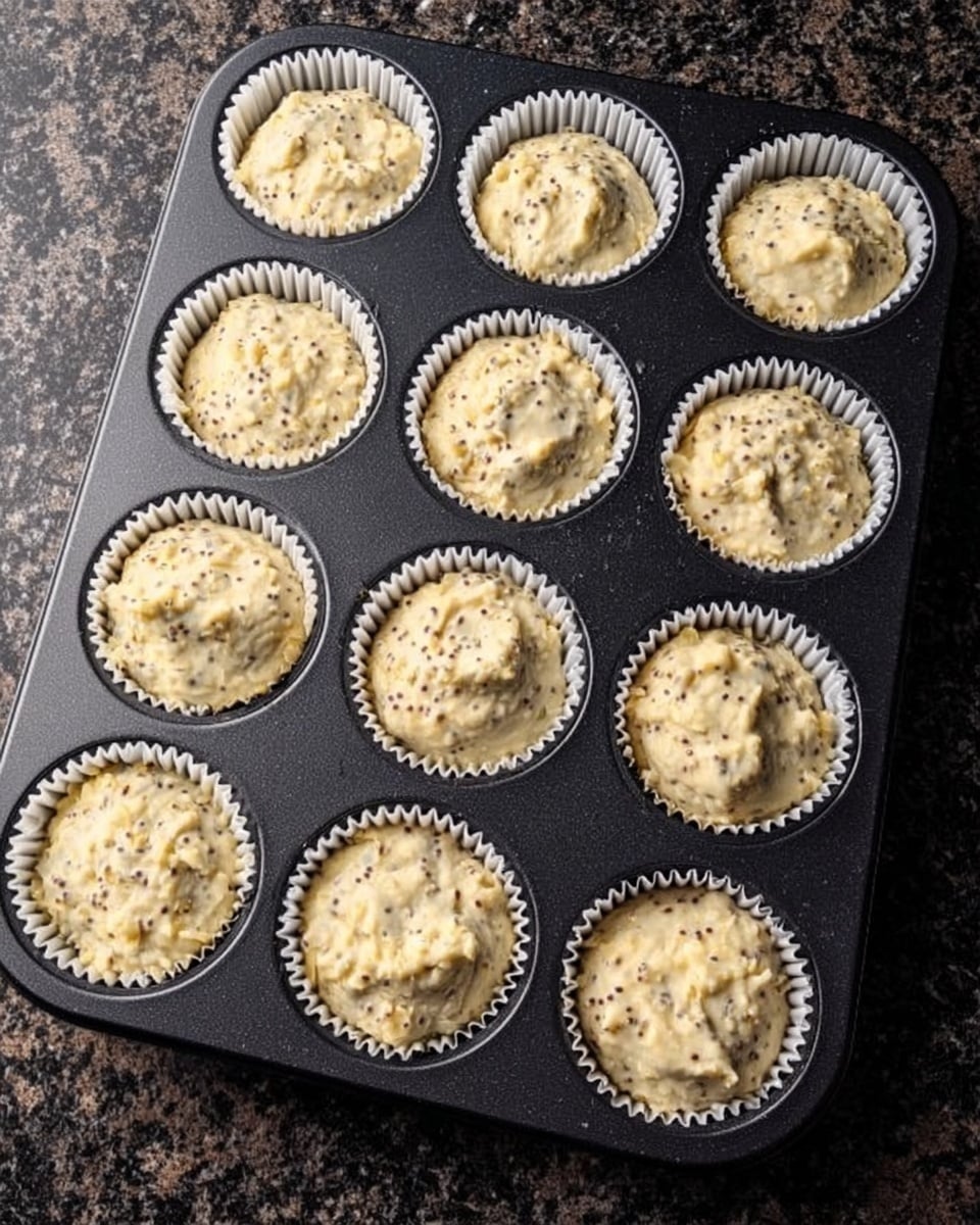 A black muffin pan holds twelve white paper liners filled with light beige batter speckled with tiny dark bits. The batter is roughly spread and slightly mounded in each liner, showing a thick texture. The pan rests on a dark granite surface with a mix of gray and black colors. photo taken with an iphone --ar 4:5 --v 7
