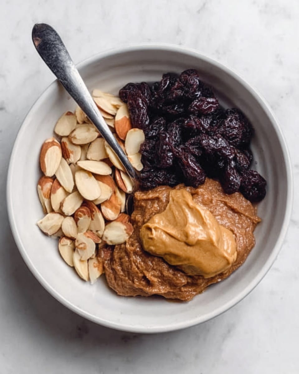 The image shows a white bowl filled with three main layers arranged side by side: on the left, sliced almonds with a light beige color and slight shine; in the middle, a thick brown paste with a soft, smooth texture; on the right, a smooth dollop of light brown peanut butter with a creamy texture. On top of the almond layer, there are dark, wrinkled pieces of dried fruit adding contrast. A silver spoon rests inside the bowl, extending upward. The bowl sits on a white marbled surface. Photo taken with an iphone --ar 4:5 --v 7