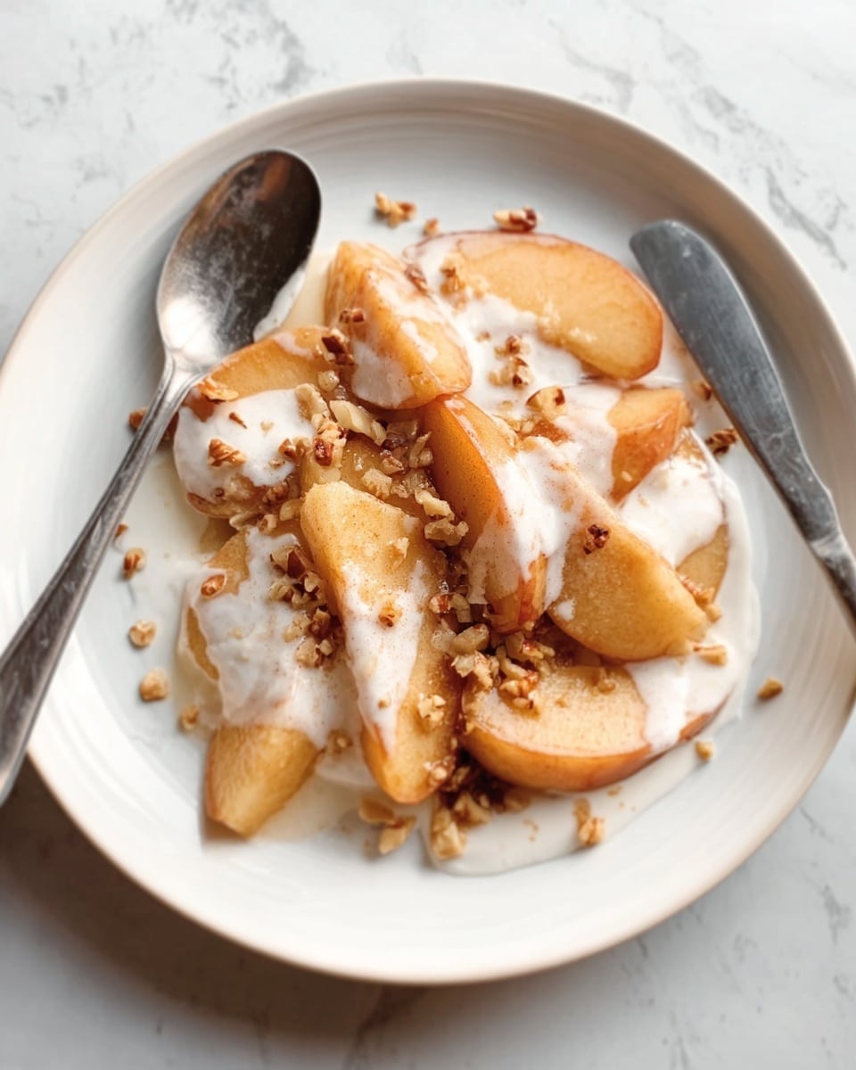 The image shows a white plate with several cooked apple slices arranged in a small pile in the center. The apples are golden brown with a soft texture and are covered with a drizzle of white cream. Scattered on top are small pieces of light brown nuts adding a crunchy look. A silver spoon and a knife are resting on the edge of the plate, which sits on a white marbled surface. The overall scene looks warm and inviting, photo taken with an iphone --ar 4:5 --v 7