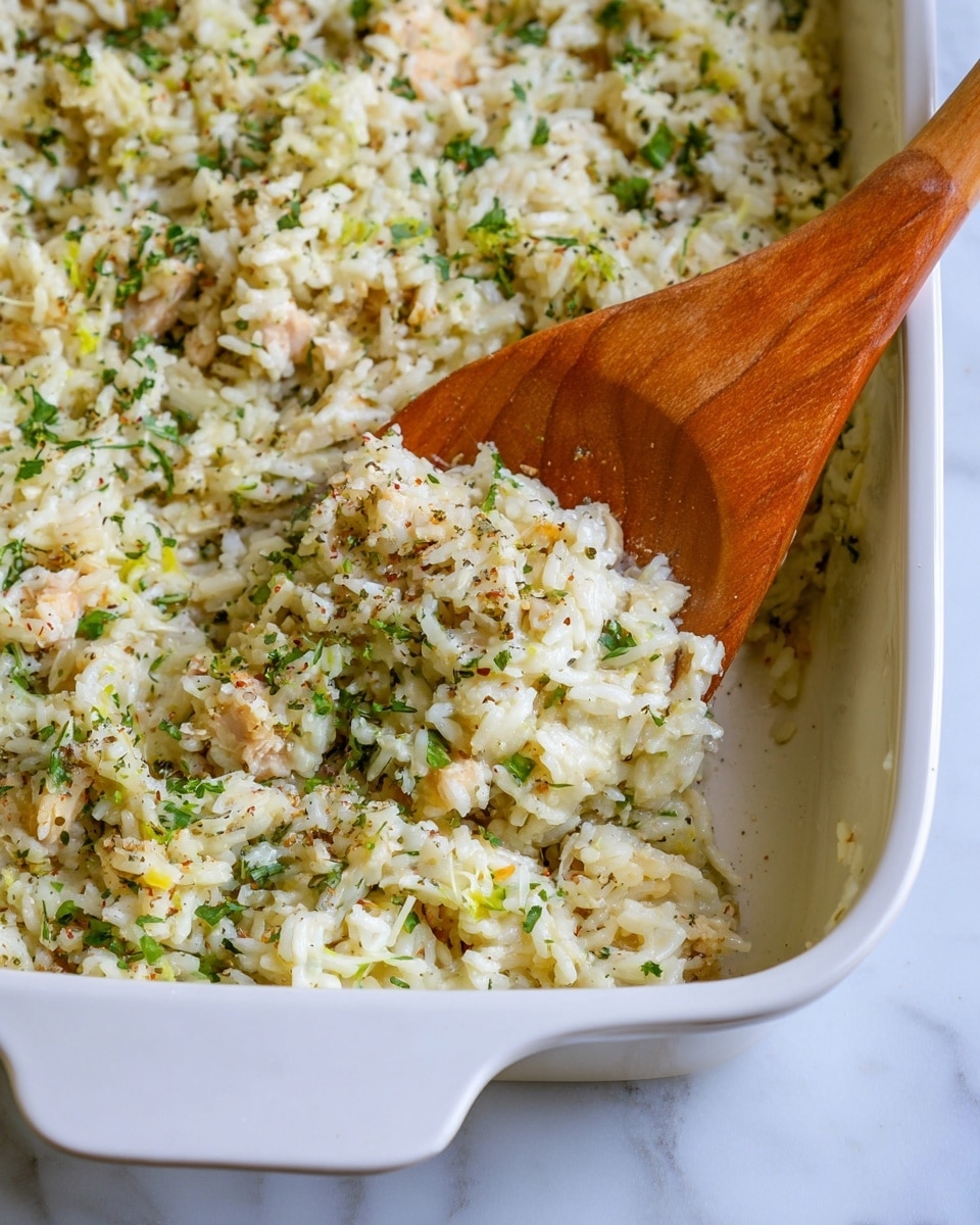 A close-up view of a white baking dish filled with a creamy rice mixture made of white rice, green herbs, small pieces of white meat, and finely grated cheese. The rice is mixed with visible green flecks of herbs and a light dusting of black pepper or spices, giving it a textured look. A wooden spoon inside the dish is scooping a portion of the rice, showing the soft, slightly clumpy texture of the dish. The baking dish is placed on a surface with a white marbled texture. Photo taken with an iphone --ar 4:5 --v 7