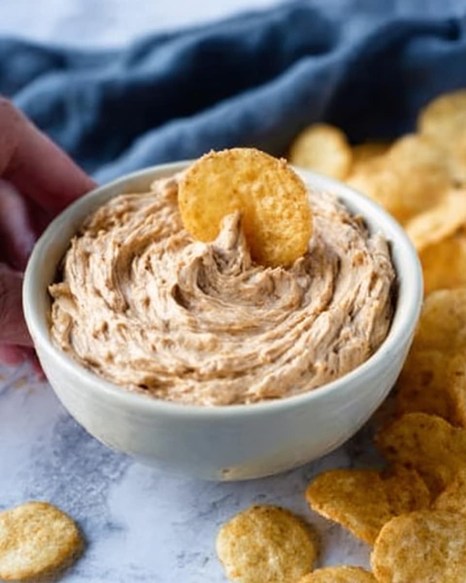 A white bowl filled with creamy light brown dip that looks soft and smooth, showing swirled textures on the surface. Three round, light beige chips stand upright in the dip, slightly textured and crispy looking. Around the bowl are scattered more chips of the same color on a white marbled surface with a soft dark blue cloth in the background. A woman's hand is holding the bowl at the edge. Photo taken with an iphone --ar 4:5 --v 7