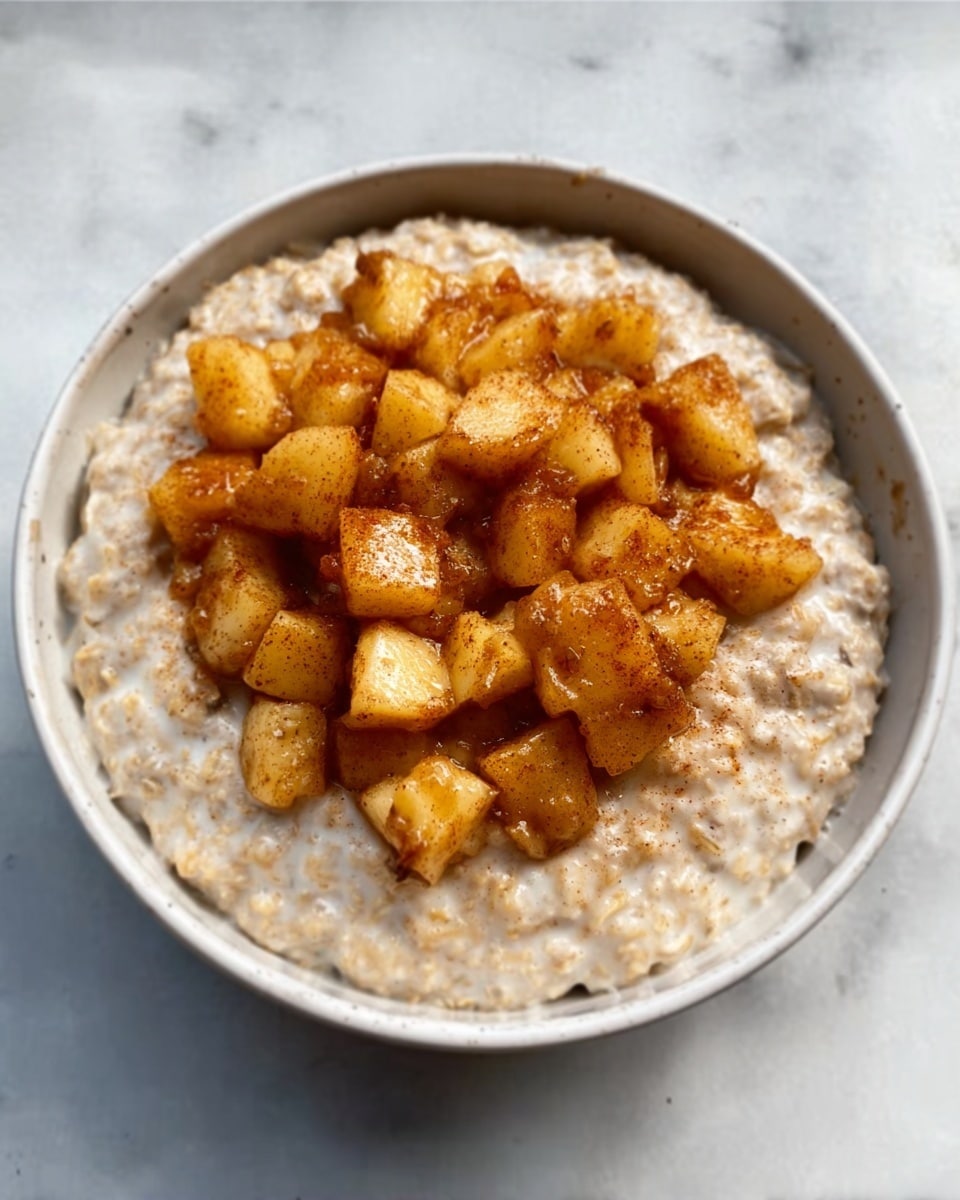 The image shows a white bowl filled with a thick layer of creamy oatmeal as the base, with small visible flakes and a smooth texture. On top, there is a generous pile of diced golden-brown cooked apples, looking soft and slightly shiny from being cooked with spices. The apple pieces are uneven in size and spread mostly in the center of the oatmeal, creating a warm color contrast between the light oatmeal and richer caramel-toned fruit. The bowl is placed on a white marbled surface. Photo taken with an iphone --ar 4:5 --v 7