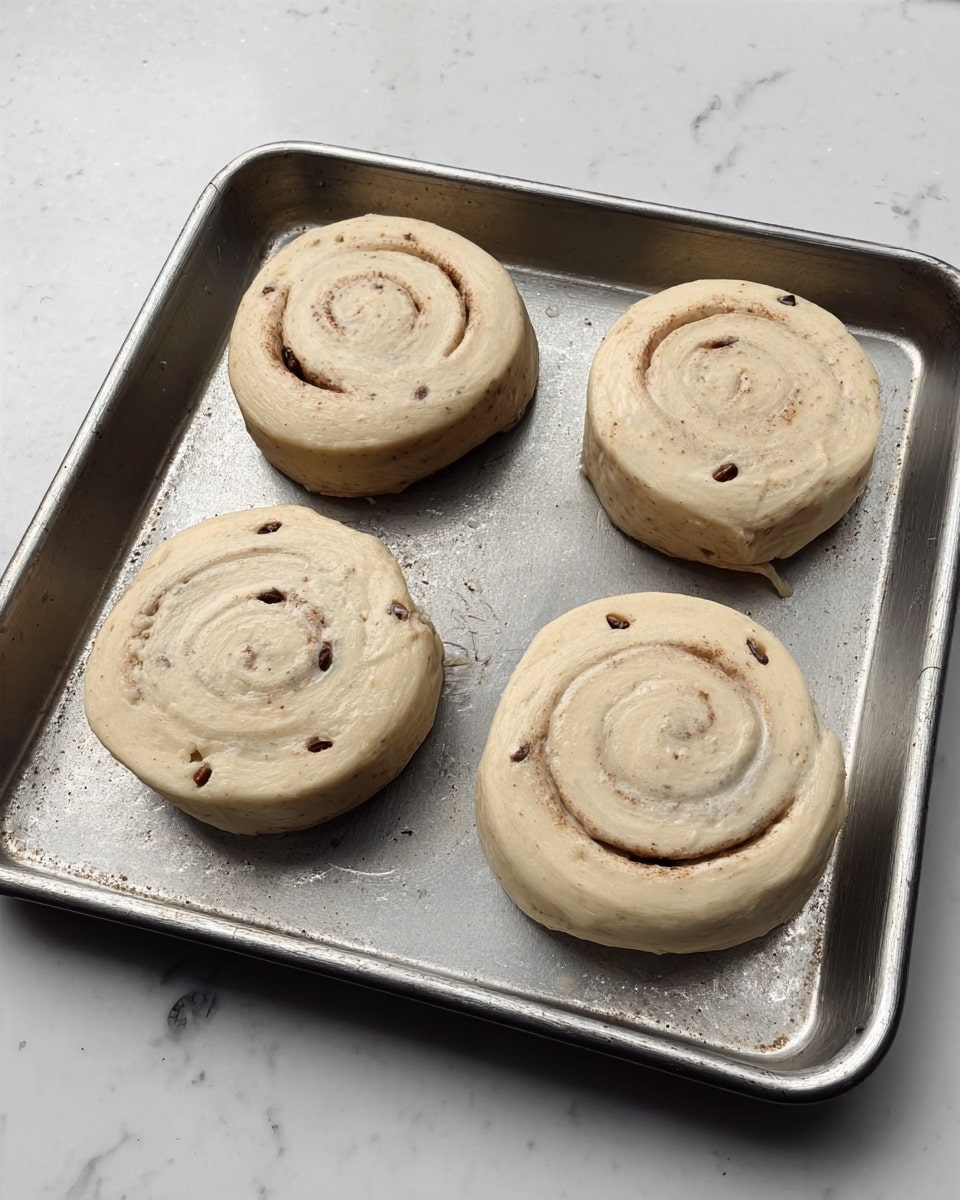 Four raw cinnamon rolls are placed on a silver metal baking tray. Each roll has a smooth, light beige dough with visible small dark spots resembling raisins or spices scattered inside. The rolls are arranged in a loose square pattern with some space between them, showing their round shape and spiral patterns clearly. The metal tray has a matte finish with slight reflections and edges visible, all set on a white marbled surface. photo taken with an iphone --ar 4:5 --v 7