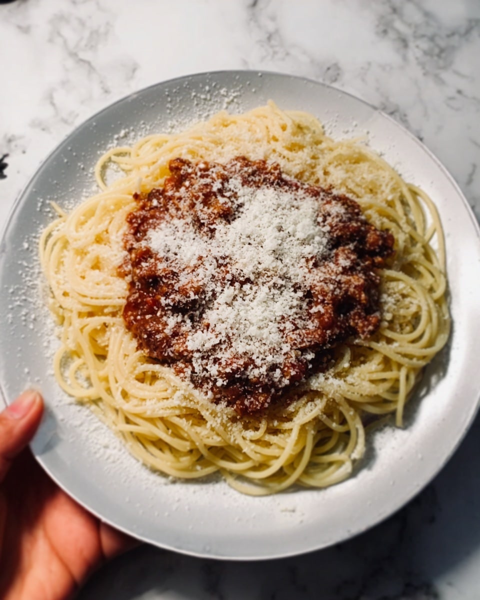 A plate of spaghetti with sauce on top placed on a white marbled surface. The spaghetti is in a loose round shape, yellowish in color with a smooth texture. On top is a layer of thick red-brown meat sauce covering the center of the pasta. Lightly sprinkled over the sauce is a fine layer of grated white cheese giving a powdery texture. A woman's hand is partially visible holding the edge of the plate. The plate itself is white with a simple round shape. Photo taken with an iphone --ar 4:5 --v 7