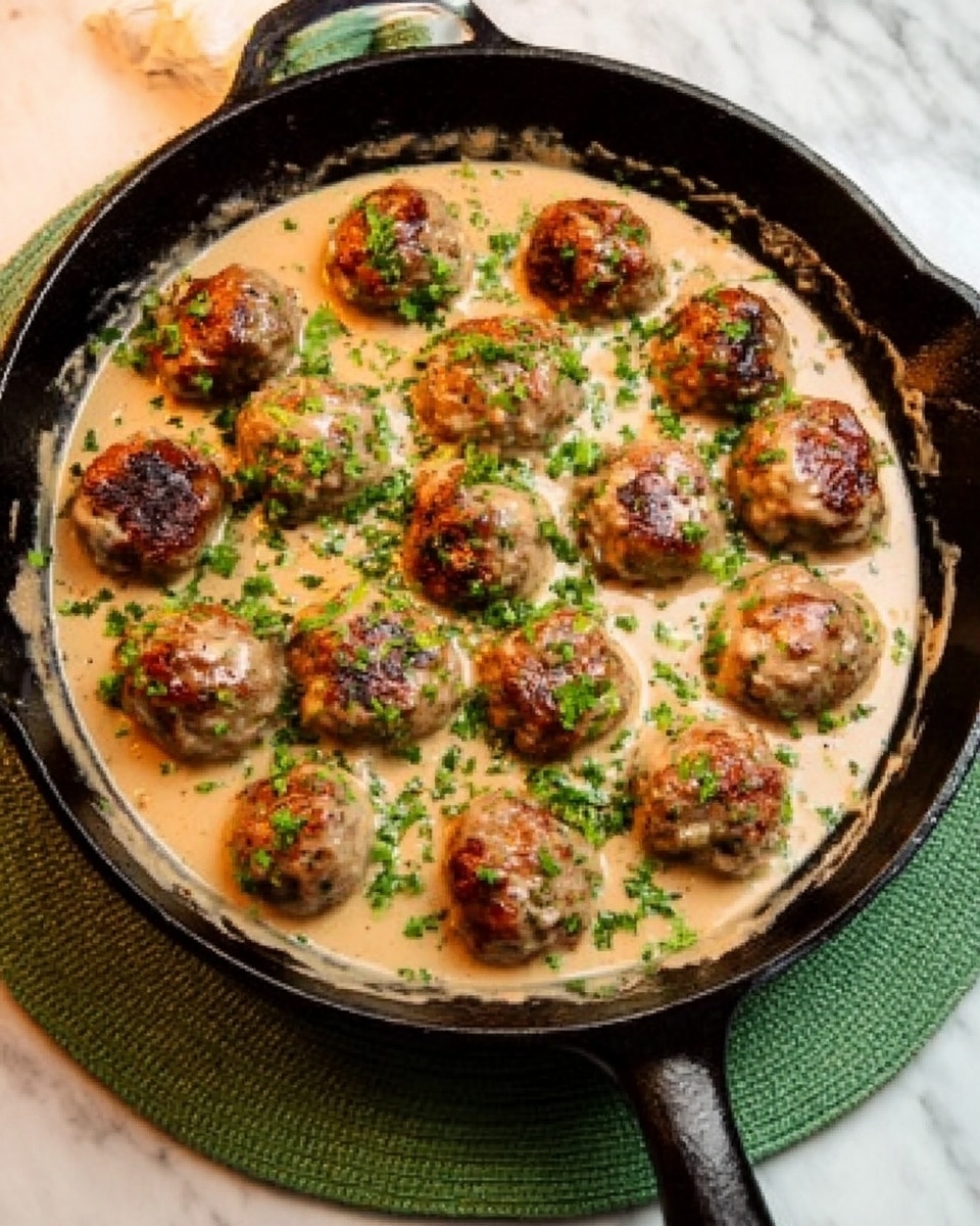 A black cast iron skillet sits on a white marbled surface with a green round cloth underneath, filled with meatballs in a creamy sauce. The meatballs are arranged in a loose circle with a few in the center, each browned and topped with chopped green herbs. The creamy sauce is light tan, smooth, and evenly spread around and under the meatballs, touching the sides of the skillet. A woman's hand is holding the skillet handle on the right side. Photo taken with an iphone --ar 4:5 --v 7
