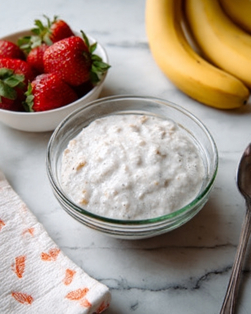 A clear glass bowl on a white marbled surface holds a thick, white mixture with small light brown pieces inside. To the left, there is a white bowl filled with red strawberries and green leaves. In the background, a few yellow bananas rest on the white marbled surface next to a silver spoon and a white napkin with small orange patterns. The overall look is fresh and simple, with soft natural lighting. photo taken with an iphone --ar 4:5 --v 7