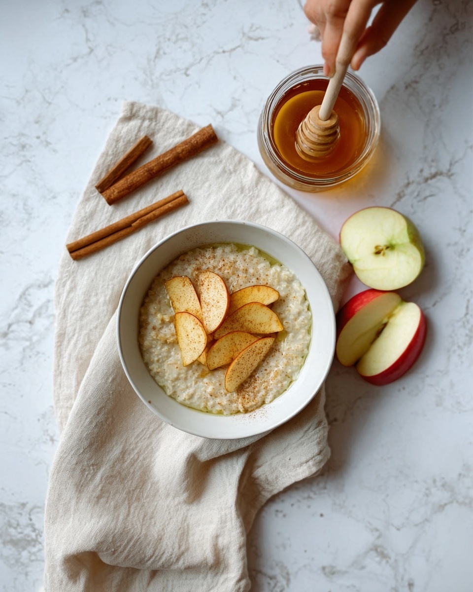 A white bowl filled with light green oatmeal topped with thin, golden brown apple slices arranged in a small pile in the center. The bowl is placed on a cream-colored cloth napkin that spreads over a white marbled surface. To the right of the bowl, there are two apple slices, one with a red skin and the other with green skin. Above the bowl, there is a small glass jar of honey with a wooden honey dipper resting inside, and two cinnamon sticks are nearby on the napkin and marble surface. A woman's hand holds the honey dipper, poised over the bowl. Photo taken with an iphone --ar 4:5 --v 7