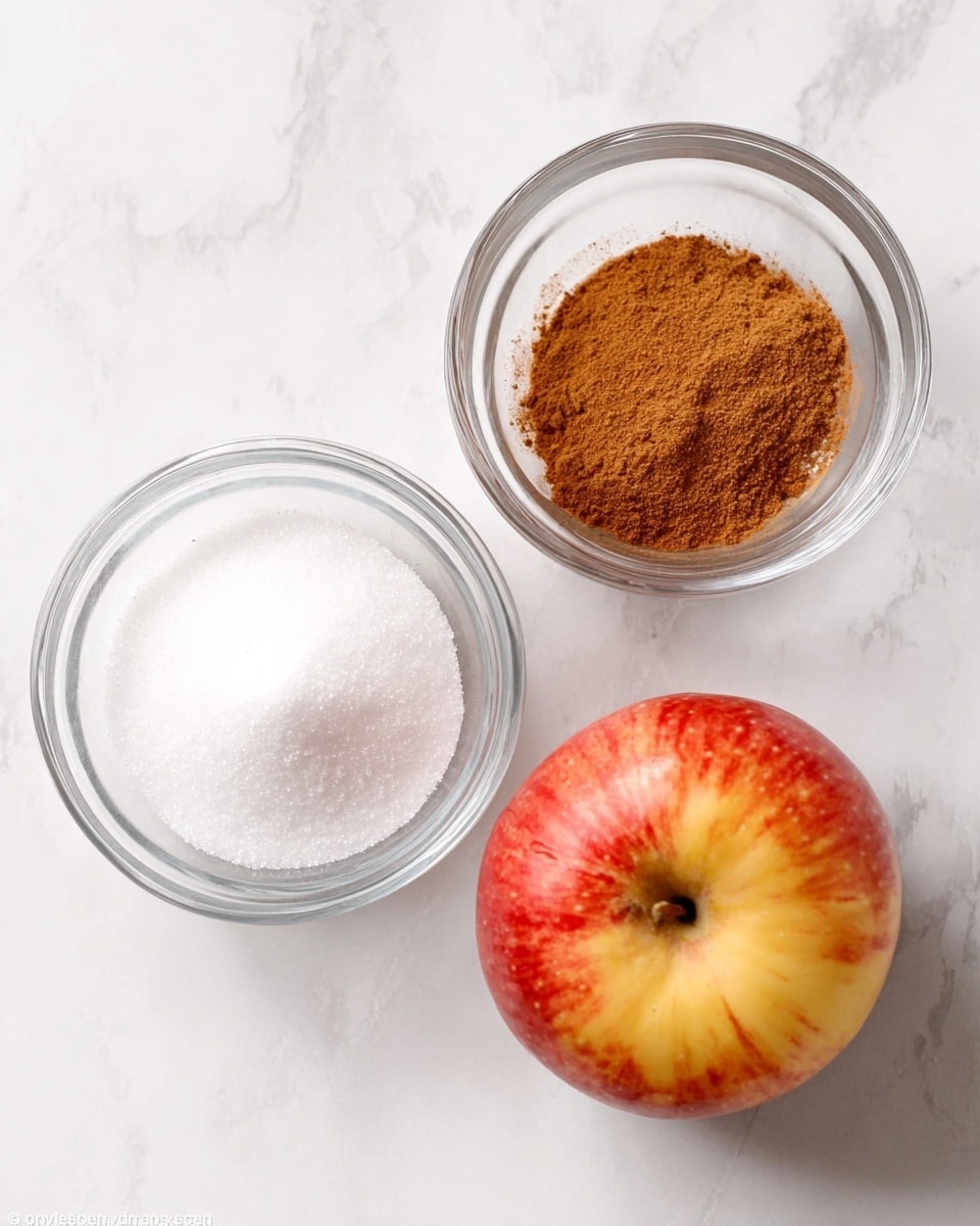 The image shows three ingredients placed on a white marbled surface: a red and yellow whole apple on the lower right, a clear glass bowl filled with white granulated sugar on the lower left, and another clear glass bowl containing a brown powder, likely cinnamon, at the top. The bowls are round and clear, allowing the textures inside to be visible. photo taken with an iphone --ar 4:5 --v 7