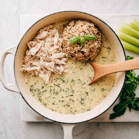 A white pan sits on a stove with a creamy light beige broth filling the bottom, speckled with green herbs. On the left side of the pan, there are shredded pieces of pale cooked chicken layered on top of the broth. On the right side, a mound of brown rice with some darker grains mixed in sits partially in the broth. A light brown wooden spoon rests inside the pan, partially submerged in the creamy broth on the right side. To the right of the pan, stalks of green celery and fresh basil leaves lie on a white marbled cutting board on a white marbled surface. photo taken with an iphone --ar 4:5 --v 7