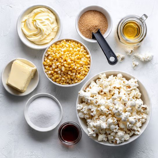 A top-down view of a white bowl full of popped popcorn with fluffy white textures and some lightly browned spots, placed in the bottom right corner on a white marbled surface. Above it, a black measuring cup filled with shiny yellow unpopped corn kernels sits near an open glass jar of white coconut oil with a black spoon inside. To the left, a white bowl holds smooth, creamy butter with a pale yellow color and swirled texture. Above it, there is a white bowl full of light brown soft brown sugar with a crumbly texture. Small white dishes with white granulated salt and white baking soda are next to the bowls. A small clear glass container with dark brown vanilla extract and a clear measuring cup filled with light golden liquid complete the arrangement. The colors are soft and natural, with a clean and tidy layout. Photo taken with an iphone --ar 4:5 --v 7