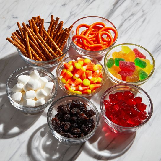 Eight clear glass bowls are arranged on a white marbled surface, each filled with different colorful snacks. Starting from the top left, there are thin brown pretzel sticks in the first bowl, bright red licorice sticks coiled in the second, and a mix of yellow, green, red, and orange gummy candies in the third. Below these, the fourth bowl contains small white marshmallows, while the fifth bowl has shiny black round candies. The sixth bowl is filled with orange, yellow, and white candy corn pieces. Finally, the seventh bowl contains dark raisins, and the eighth bowl holds small red candies. All bowls are round with smooth edges, and daylight casts soft shadows around them. Photo taken with an iphone --ar 4:5 --v 7