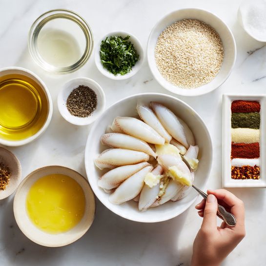 The image shows a white bowl in the center filled with raw, small, whole squids arranged neatly. Around it, there are several smaller white bowls and dishes placed on a white marble surface. The smaller bowls contain different ingredients: clear liquid, chopped green herbs, black pepper, yellow grated zest, and minced garlic. A round white dish contains sesame seeds. There is also a clear bowl with a yellow liquid oil, a small white bowl with salt, and a small square white dish holds three rows of different spices in shades of red, green, and brown. In the bottom left, a clear bowl contains a thick yellow butter-like substance. A woman's hand is visible, holding a spoon over the butter-like mixture. The whole setting has soft, natural light. Photo taken with an iphone --ar 4:5 --v 7