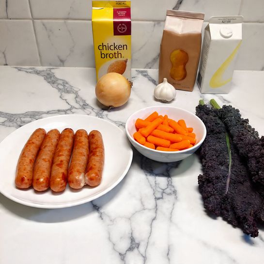 A white plate holds six brown sausages arranged neatly on the left side. Next to the plate, there is a small white bowl filled with bright orange baby carrots. Behind the bowl, a yellow onion and a single white garlic bulb rest on a white marbled surface. Behind these items, three cartons are visible: a yellow one labeled 