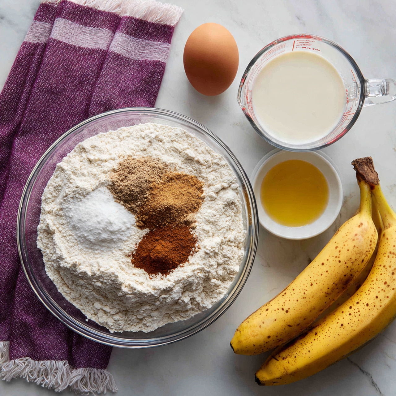 A clear glass bowl in the top right holds several layers of dry ingredients: a large amount of white flour covering most of the bowl, with a small pile of brown cinnamon and another smaller pile of lighter brown spice on top towards the upper left side; below is a fine white powder, likely baking powder or sugar. To the left of the bowl, a purple and white striped cloth with fringed edges lies on a white marbled surface. Near the middle left of the image sits a single brown egg. Below it are two small white bowls: one with dark brown vanilla extract and another filled with a yellow liquid, possibly melted butter or oil. In the middle right, a clear glass measuring cup contains a white liquid, which looks like milk. At the bottom right, two yellow bananas with small brown spots lie side by side. The photo was taken with an iphone --ar 4:5 --v 7