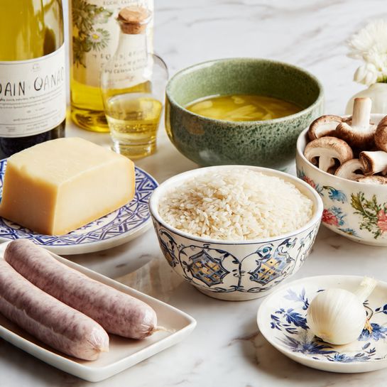 Mushroom, Fennel and Sausage Risotto Recipe 3 The image shows an arrangement of cooking ingredients on a white marbled surface. At the front is a white rectangular plate with two raw sausages, light purple in color with smooth texture. To the right, a white round bowl with floral blue patterns is filled with raw Arborio rice, small and white. Behind it, a green ceramic bowl with a rustic look holds golden-yellow broth. On the left side, a white square plate with blue geometric patterns holds a block of pale yellow cheese with a hard texture. Next to it is a white round bowl with a dark blue crisscross pattern, filled with sliced brown mushrooms, showing a rough texture. To the right of the mushrooms sits an unpeeled onion on a small round white dish with blue flower designs. Behind all, there is a bottle of white wine with a cream label and an olive oil bottle with a black label. Photo taken with an iphone --ar 4:5 --v 7