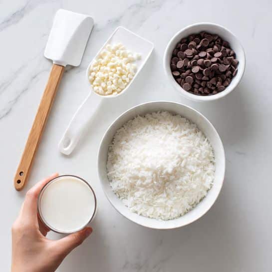 The image shows three white bowls on a white marbled surface. The largest bowl in the center is full of small white chips with a smooth, slightly rounded texture. In the top right, a smaller bowl contains dark brown chocolate chips, which are round and glossy. Above the largest bowl on the left side, there is a white spatula with a wooden handle resting on the surface. A woman's hand is holding a clear glass filled with white milk, positioned near the bottom center over the large bowl of white chips. photo taken with an iphone --ar 4:5 --v 7