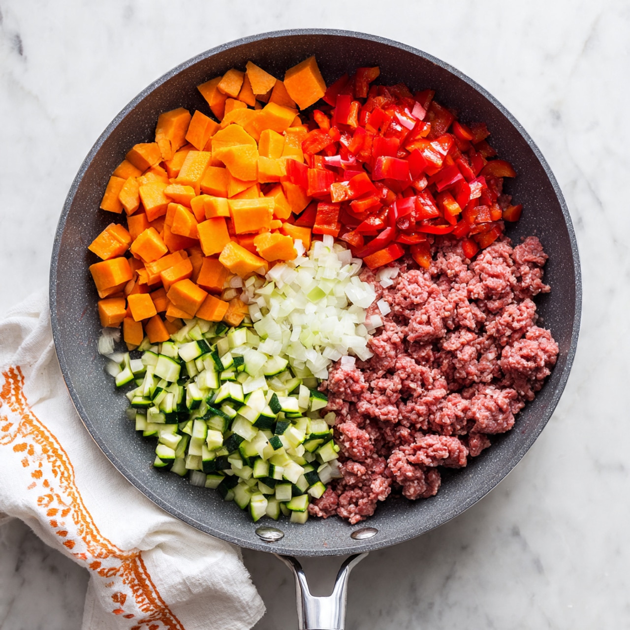 A dark gray frying pan on a white marbled surface holds six different ingredients neatly separated in layers: a large pile of small orange cubes of sweet potato at the top left, next to bright red chopped bell peppers in the middle, followed by light green chopped zucchini pieces on the bottom left, minced garlic below the peppers, finely chopped white onions next to the garlic, and raw ground meat in a wavy pattern filling the entire right side of the pan. A woman's hand is holding the silver handle of the pan, which rests on a white cloth with an orange pattern. photo taken with an iphone --ar 4:5 --v 7