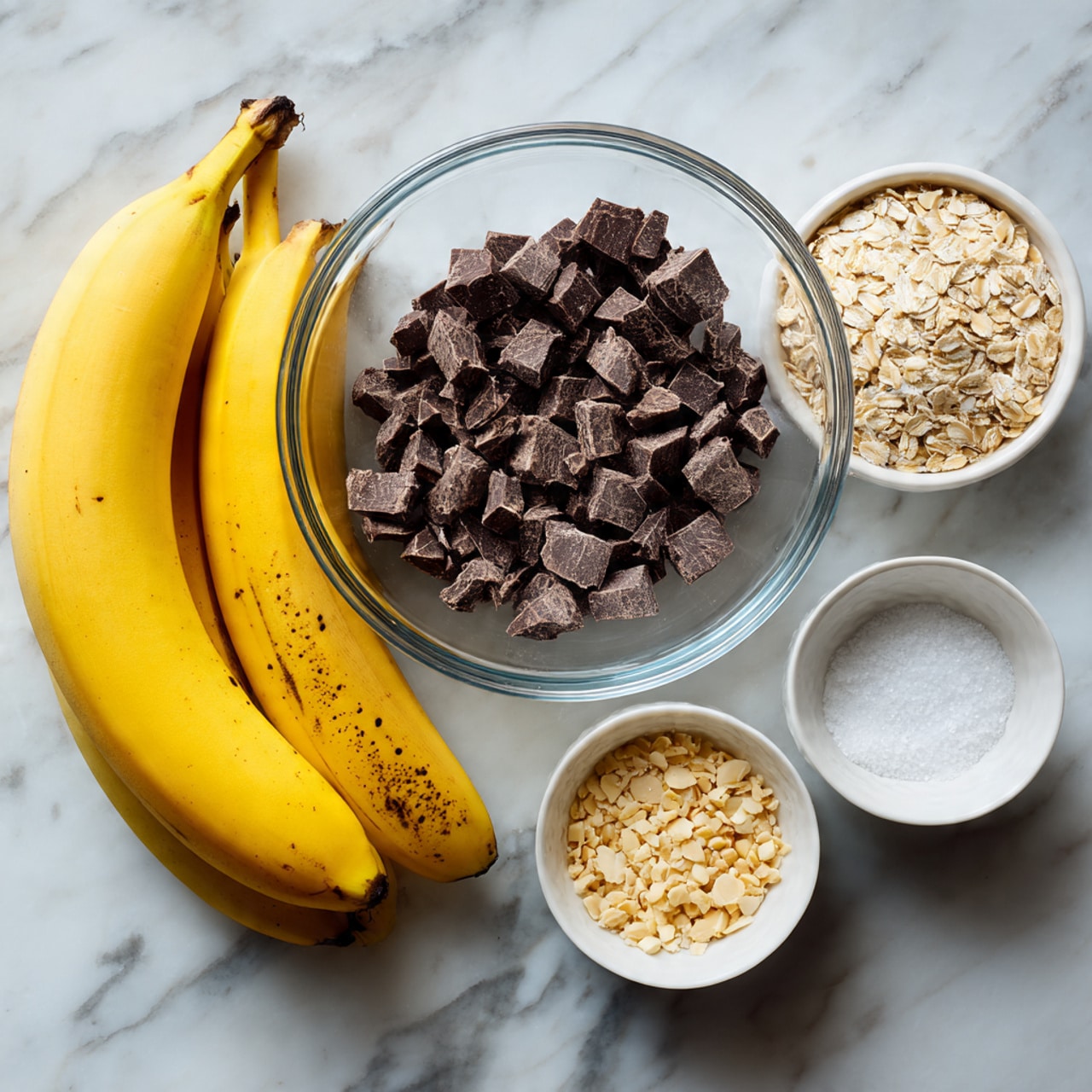 A clear glass bowl filled with small dark chocolate chunks sits near two ripe yellow bananas with brown spots, placed on a white marbled surface. Around the glass bowl, three small white bowls hold different ingredients: one with light brown oats, another with crushed pale nuts, and the last with fine white salt. The arrangement is neat with all bowls spaced evenly. Photo taken with an iphone --ar 4:5 --v 7