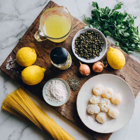 A dark wooden cutting board on a white marbled surface holds scattered ingredients: a clear glass measuring cup with light yellow broth on the top left, a white bowl full of dark green capers above it, a small bottle with a black lid in the center, two bright yellow lemons slightly to the right, and a bunch of green parsley with stems visible to the top right. Below them are two brown shallots near a whole garlic bulb. A clear glass bowl with white flour sits near the center, while a mound of creamy white scallops fills a round white plate to the bottom right. On the bottom left, uncooked yellow spaghetti is upright in a clear glass. photo taken with an iphone --ar 4:5 --v 7