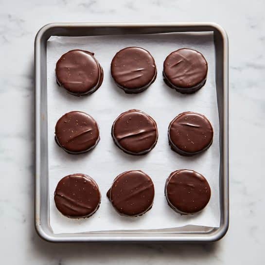 The image shows a gray baking tray lined with white parchment paper on a white marbled surface. On the tray, there are twelve round chocolate-covered treats arranged in three rows and four columns. Each treat is fully coated in smooth milk chocolate, with some slight unevenness and small drips on the edges. The chocolate has a shiny texture with soft reflections, and a few small cracks or lines can be seen on the surface of some pieces. The overall look is neat but homemade, with some chocolate spreading slightly onto the parchment paper around the treats. photo taken with an iphone --ar 4:5 --v 7