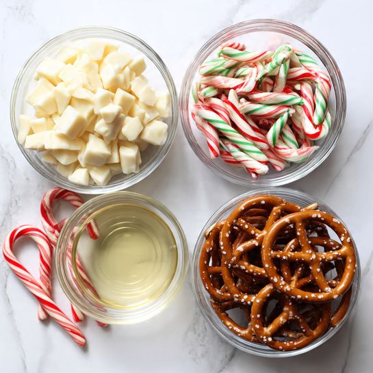 The image shows four clear glass bowls on a white marbled surface. The top left bowl holds many small, white chocolate chunks piled high with a smooth, slightly powdery texture. The top right bowl is filled with many small candy canes striped in red, green, and white, all of them curved at one end and packed closely together. Below these two bowls, a smaller bowl in the center contains a small amount of a clear, pale yellow liquid. The bottom bowl is filled with many glossy, brown pretzels stacked loosely, showing their twisted shapes and small white salt crystals. Around the bowls, a few candy canes are scattered on the white marbled surface. Photo taken with an iphone --ar 4:5 --v 7