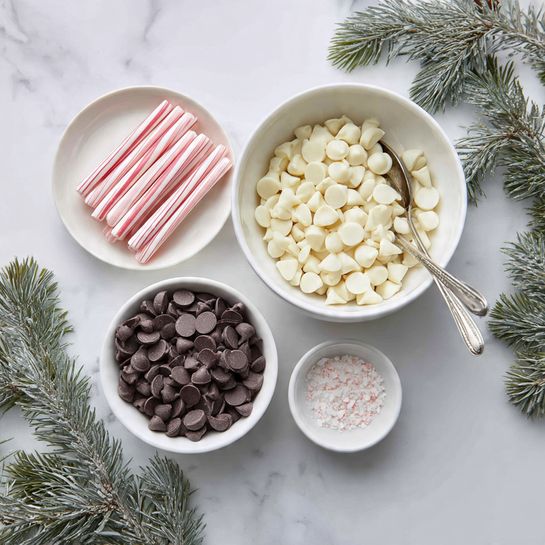 The image shows three white dishes on a white marbled surface. The top right dish is a large white bowl filled with small white pieces that look like white chocolate chips. A silver spoon rests inside this bowl. To the left is a small white plate with pink and white striped candy sticks stacked neatly. Below both dishes is another white bowl filled with dark brown chocolate chips, also with a silver spoon inside. A tiny white bowl to the right contains a small amount of white granules. Some green pine branches are placed around the dishes for decoration. Photo taken with an iphone --ar 4:5 --v 7