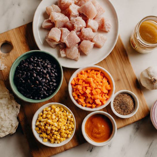 The image shows a white plate with light pink cooked chicken pieces on it, placed on the top left of a wooden cutting board. On the cutting board, there are several small white bowls and piles of ingredients: a bright green bowl with black beans on the bottom left, a small white bowl with yellow corn next to it, chopped white onion and bright orange chopped carrots in the center, a small bowl with a red sauce near the bottom edge, and a tiny bowl with a brown spice mix at the top right. There is also a glass jar with a light brown liquid near the top right corner of the board. All of this is set on a white marbled surface. photo taken with an iphone --ar 4:5 --v 7