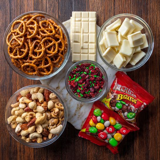 The image shows five items placed on a dark wooden surface with a white marbled texture. There is a clear glass bowl filled with light brown pretzels, a smaller clear glass bowl full of mixed roasted peanuts with some red skins, and a white chocolate bar with squares visible. Next to these, a round container has small red and green sprinkles, and there are two orange bags of snacks: one is a red bag of Bugles snack and the other is a Christmas-themed M&M's candy bag with red, green, and white colors. Photo taken with an iphone --ar 4:5 --v 7