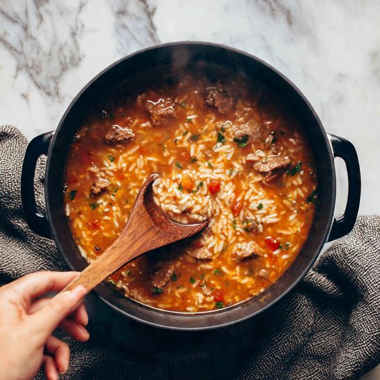A black pot filled with a thick stew that has a mix of rich orange and red colors with pieces of cooked rice, small green leaves, and chunks of light brown meat scattered throughout. A wooden spoon is stirring the stew, held by a woman's hand, with steam lightly rising, showing the hot texture. The pot sits on a white marbled surface with a textured gray cloth underneath. photo taken with an iphone --ar 4:5 --v 7