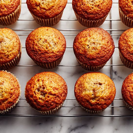 The image shows a cooling rack holding twelve golden brown muffins arranged in neat rows. Each muffin has a slightly domed top with a light, crumbly texture, and small cracks visible on the surface, indicating softness inside. The cooling rack is sitting on a white marbled surface, and the muffins sit in paper liners that are light beige and tightly wrapped around each muffin base. Photo taken with an iphone --ar 4:5 --v 7