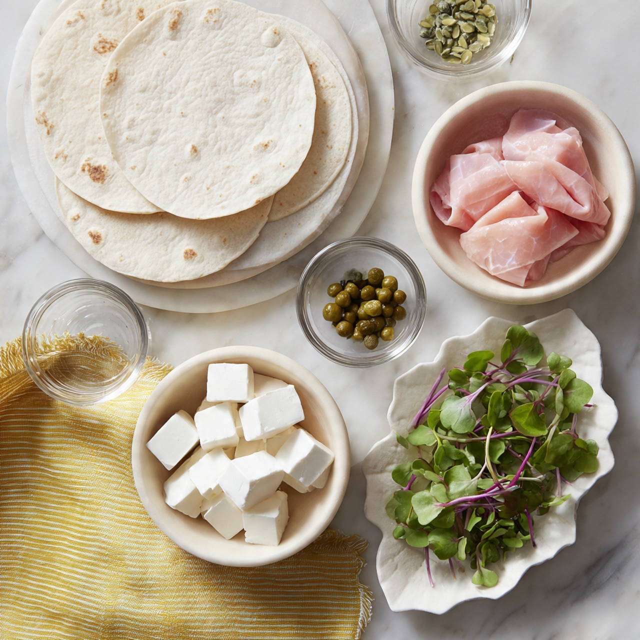 The image shows several small white bowls and plates arranged on a white marbled surface with a yellow striped cloth nearby. One white circular plate holds round white tortillas, while a small white bowl contains soft white cheese cubes. Another small white bowl is filled with pinkish sliced smoked salmon. A small glass bowl holds green capers, and a white leaf-shaped dish has fresh green microgreens with tiny purple leaves. The setup looks fresh and colorful, showing ingredients ready for making wraps or rolls. photo taken with an iphone --ar 4:5 --v 7