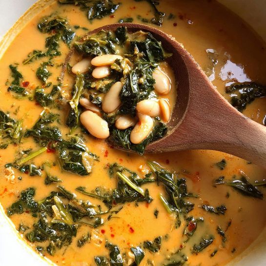 A close-up view of a creamy orange soup with visible pieces of cooked green leafy vegetables mixed evenly throughout. On top, there are small white beans clustered near the edge of the soup, slightly submerged. A wooden spoon is resting inside the white pot, scooping up part of the soup. The background surface is a white marbled texture. Photo taken with an iphone --ar 4:5 --v 7
