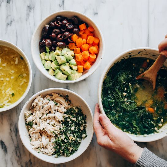 Three white bowls sit on a white marbled surface. The first bowl shows three layers: a top layer of black olives, a middle layer of creamy yellow liquid, and a layer with small light beige beans. The second bowl contains chopped green avocado, white chopped onions, and bright orange carrot slices arranged side by side. The third bowl has chopped green herbs like dill, mixed with smaller bits of orange carrot and some seeds sprinkled on top. In the next photo, two white bowls are side by side on the white marbled surface. The left bowl is filled with the mixed chopped herbs, carrots, and seeds from before, while the right bowl has a yellowish thick soup with pieces of white cannellini beans on top. The final image shows one white bowl filled with dark green cooked spinach on the white marbled surface and another white bowl with the mixed soup stirred, showing soft orange carrot slices and spinach floating. A woman's hand holding a wooden spoon is partly visible stirring the soup. Photo taken with an iphone --ar 4:5 --v 7