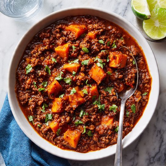 A white bowl filled with thick chili that has layers of dark brown ground meat, bright orange chunks of sweet potatoes, and small bits of green cilantro sprinkled on top. The chili looks rich and saucy with a deep red color. A silver spoon is resting inside the bowl, scooping up some of the chili. Next to the bowl are two lime wedges on a white marbled surface and a clear glass of water. A blue cloth napkin is on the side. The photo has a cozy, homemade feel. photo taken with an iphone --ar 4:5 --v 7